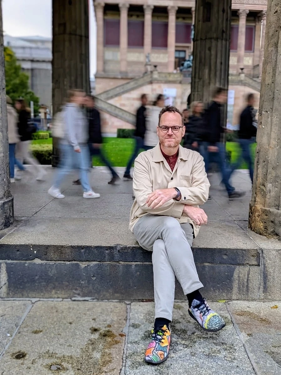 Eyal seated on stone steps in a light jacket, with blurred visitors passing behind him